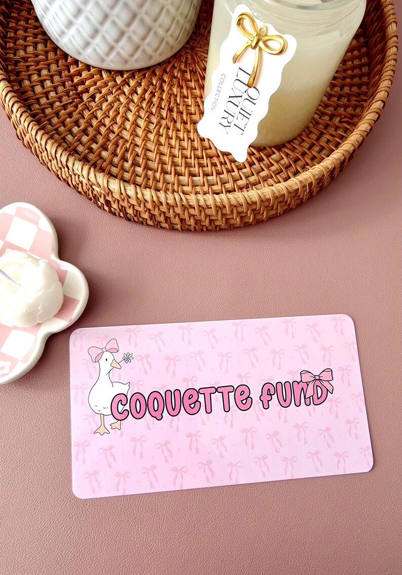 May include: A pink rectangular card with the words "COQUETTE FUND" in pink bubble letters, and a cartoon goose with a pink bow. The background has a pattern of pink bows. A woven basket and a candle are in the background.