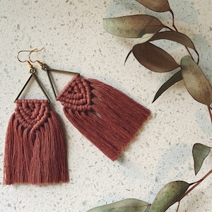 May include: A pair of handmade macrame earrings. Each earring features a triangular brass frame with a woven, reddish-brown tassel. The earrings are displayed on a speckled white surface, with a sprig of dried leaves in the background.