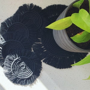 May include: Set of five black crocheted coasters with fringe. The coasters are arranged in a semi-circle with a potted plant in the background.