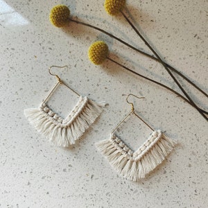 May include: A pair of gold square earrings with white macrame fringe. The earrings are on a white background with yellow flowers.