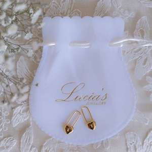 Peut inclure: Une paire de boucles d'oreilles en forme de fermoir de sécurité dorées avec des extrémités en forme de cœur, présentées sur une pochette en velours blanc avec le texte "Lucia's Jewellery".