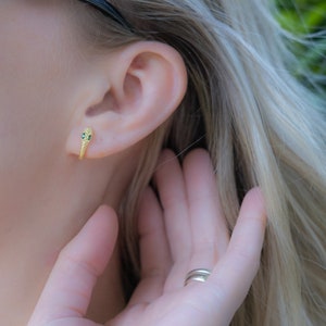 May include: A close-up of a person's ear with a gold snake earring with green eyes. The earring is small and delicate.