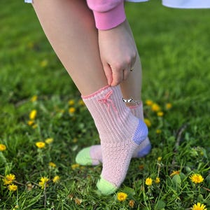 Op de afbeelding: Een paar roze sokken met een lichtroze strik aan de bovenkant. De hiel is lichtpaars en de teen is lichtgroen. De sokken zijn geribbeld en lijken handgemaakt. De persoon staat in een grasveld met gele bloemen.