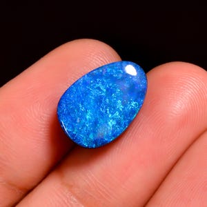 May include: A vibrant blue opal gemstone with a pear-like shape. The stone displays a mix of bright and dark blue hues, with a glossy, polished surface. The opal is held in a hand against a dark background.