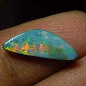 May include: A polished, triangular-shaped opal gemstone featuring a smooth, domed surface. The stone displays a vibrant play-of-color with iridescent flashes of bright orange, yellow, and green against a translucent, light blue base.