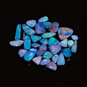 May include: A collection of loose, polished opals in various irregular shapes and sizes. These gemstones display vibrant flashes of blue, green, and red iridescence against a dark, semi-translucent base, creating a colorful, shimmering display.