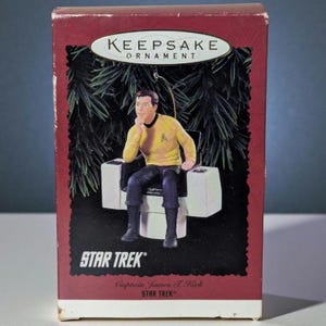 May include: A Star Trek Captain James T. Kirk Keepsake Ornament. The ornament features Captain Kirk in his yellow uniform, seated in a white chair. The box is red with the Star Trek logo and the words "Keepsake Ornament."