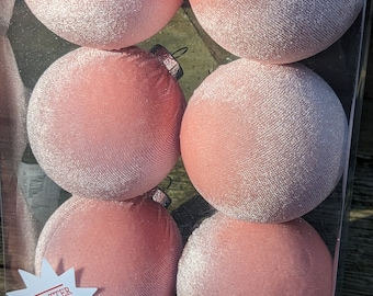May include: Six pink velvet Christmas ornaments in a clear plastic package. The ornaments are round and have a soft, textured appearance. A white star-shaped sticker with the words "SHATTER PROOF" is visible.