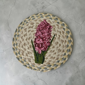 May include: Round woven coaster with a floral design. The coaster features a central image of a pink hyacinth flower with green leaves. The border is a mix of beige, gray, and yellow braided rope.