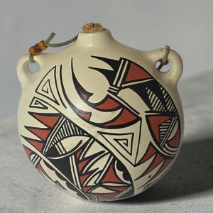 May include: Cream-colored, round ceramic flask with a cork stopper and a leather cord. The flask is adorned with geometric designs in black, red, and white. It has two small handles and is likely handmade.