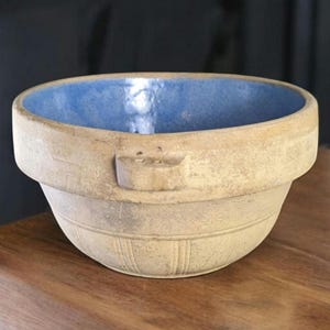 May include: A round ceramic bowl with a matte beige exterior and a smooth, vibrant blue glazed interior. The bowl features a thick rim, a small lug handle on the side, and subtle vertical embossed lines decorating the lower exterior surface.