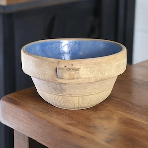 May include: A vintage blue and beige ceramic mixing bowl with a handle. The bowl has a blue glaze on the inside and a beige glaze on the outside. The bowl is sitting on a wooden table.