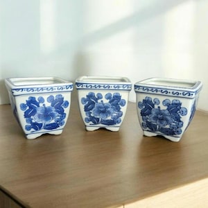 May include: Three square ceramic planters with a white base and blue floral designs. The planters have a slightly flared shape and are arranged on a wooden surface. Each planter features a different floral pattern, adding visual interest.