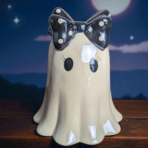 May include: A ceramic ghost figurine with a black bow featuring white polka dots. The ghost has large black eyes and delicate eyelashes. The figurine is off-white and has a rounded bottom.