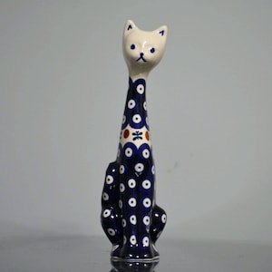 May include: A ceramic cat figurine with a tall, slender neck and a cream-colored head. The body is decorated with a dark blue glaze featuring white circular patterns, small brown accents, and a central butterfly motif on the chest area.