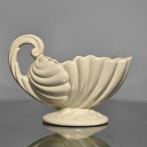 May include: A cream-colored ceramic gravy boat featuring a scalloped shell-like design with a curved handle and a pedestal base. The glossy finish highlights the vertical ridges that extend from the base to the scalloped rim.