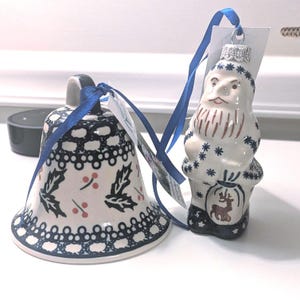 May include: Two ceramic Christmas ornaments: a bell and a Santa Claus figure. The bell is white with a black lace design, holly leaves, and red berries. The Santa ornament is white with blue snowflake details and a brown beard. Both have blue ribbons.