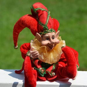 May include: A jester doll wearing a red velvet outfit with green trim and gold accents. The doll features a pointed hat with a small bell, a ruffled gold collar, and pointed shoes with bells. The face has large ears and a smiling expression.