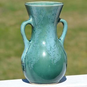 May include: A teal-colored ceramic vase with two curved handles. The vase has a bulbous body and a flared rim. The surface has a crackled texture with lighter blue-green accents, creating a unique, handcrafted appearance. The vase is approximately 8 inches tall.