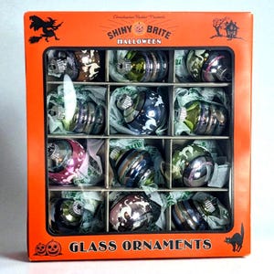 May include: A boxed set of twelve Shiny Brite Halloween glass ornaments in an orange package. The box features a witch, a haunted house, and a black cat illustration. Each round ornament has a metallic finish with various seasonal patterns.