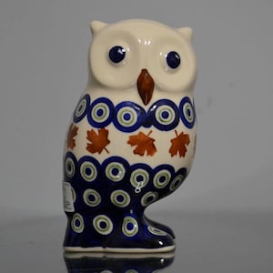 May include: A ceramic owl figurine featuring a cream-colored body with a hand-painted pattern of dark blue and light green concentric circles, interspersed with small orange maple leaf motifs, and a solid brown beak with dark blue circular eyes.