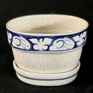 May include: A white ceramic planter with a blue decorative band around the rim and a matching saucer. The planter has a crackle glaze finish. The blue band features a repeating floral design. The planter is suitable for indoor or outdoor use.