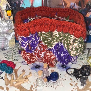 May include: A crocheted dice bag with a red, green, purple, and white pattern. The bag is filled with dice and there are several small bottles and a metal keychain with a dice charm on the table.