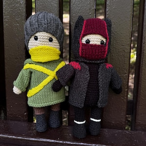 Peut inclure: Deux poupées tricotées à la main. L'une porte un bonnet gris, un pull vert, une écharpe jaune et des bottes noires. L'autre est vêtue d'une tenue noire de style ninja avec un masque et des accents rouges. Les deux poupées ont des yeux noirs.