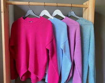 May include: Four sweaters hanging on a wooden rack. The sweaters are in different colors: pink, light blue, lavender, and light green.