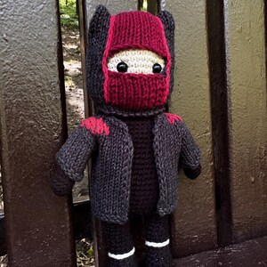 Könnte beinhalten: Handgefertigte Ninja-Puppe aus Häkelarbeit. Die Puppe hat eine rot-schwarze Maske, eine schwarze Jacke mit roten Akzenten und eine schwarze Hose mit weißen Streifen. Die Puppe sitzt auf einer braunen Bank.
