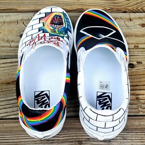 May include: A pair of custom Vans slip-on shoes with a black and white brick wall design on one shoe and a black background with a rainbow and a prism on the other shoe. The shoes have the word "Vans" printed on the side.