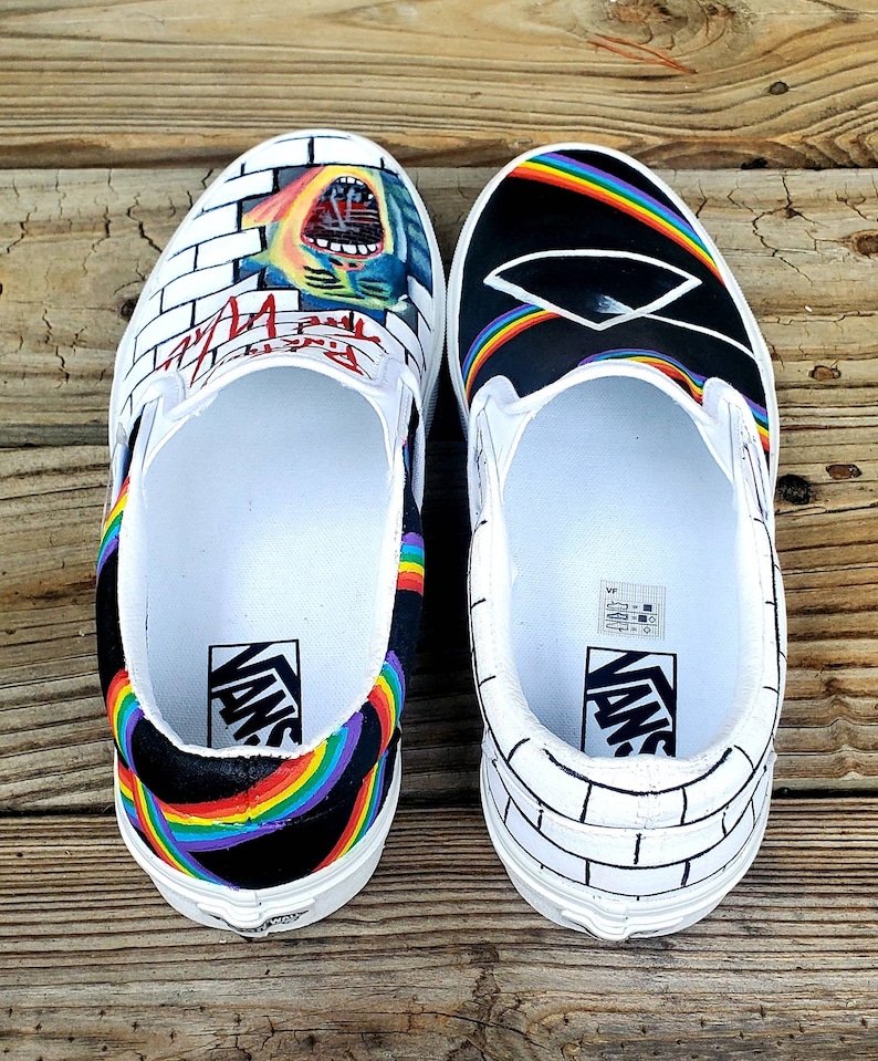May include: A pair of custom Vans slip-on shoes with a black and white brick wall design on one shoe and a black background with a rainbow and a prism on the other shoe. The shoes have the word "Vans" printed on the side.