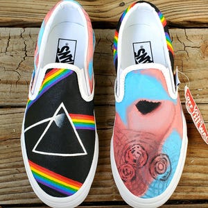 May include: A pair of black canvas slip-on Vans shoes with custom painted designs. One shoe features a rainbow and a black triangle with a white outline. The other shoe features a pink and blue abstract design with black circles. The shoes have white soles and the Vans logo is visible on the back of each shoe.