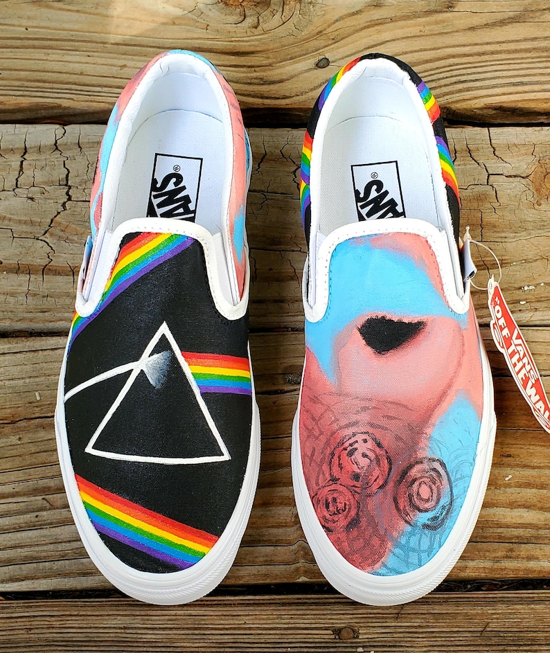 May include: A pair of black canvas slip-on Vans shoes with custom painted designs. One shoe features a rainbow and a black triangle with a white outline. The other shoe features a pink and blue abstract design with black circles. The shoes have white soles and the Vans logo is visible on the back of each shoe.