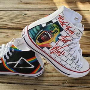 May include: A pair of high-top sneakers with custom artwork. One shoe features a black and white brick wall with the words "Pink Floyd The Wall" and a graphic of a man's face with an open mouth. The other shoe features a black background with a rainbow stripe and a black triangle with a white outline.