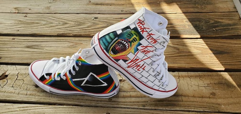May include: A pair of high-top sneakers with custom artwork. One shoe features a black and white brick wall with the words "Pink Floyd The Wall" and a graphic of a man's face with an open mouth. The other shoe features a black background with a rainbow stripe and a black triangle with a white outline.