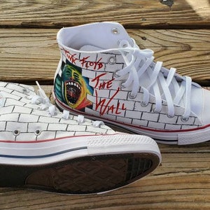 May include: A pair of white high-top sneakers with a brick wall design and a Pink Floyd "The Wall" graphic on one shoe. The shoes have white laces and a red stripe around the sole.