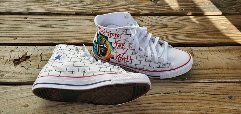 May include: A pair of white high-top sneakers with a brick wall design and a Pink Floyd "The Wall" graphic on one shoe. The shoes have white laces and a red stripe around the sole.