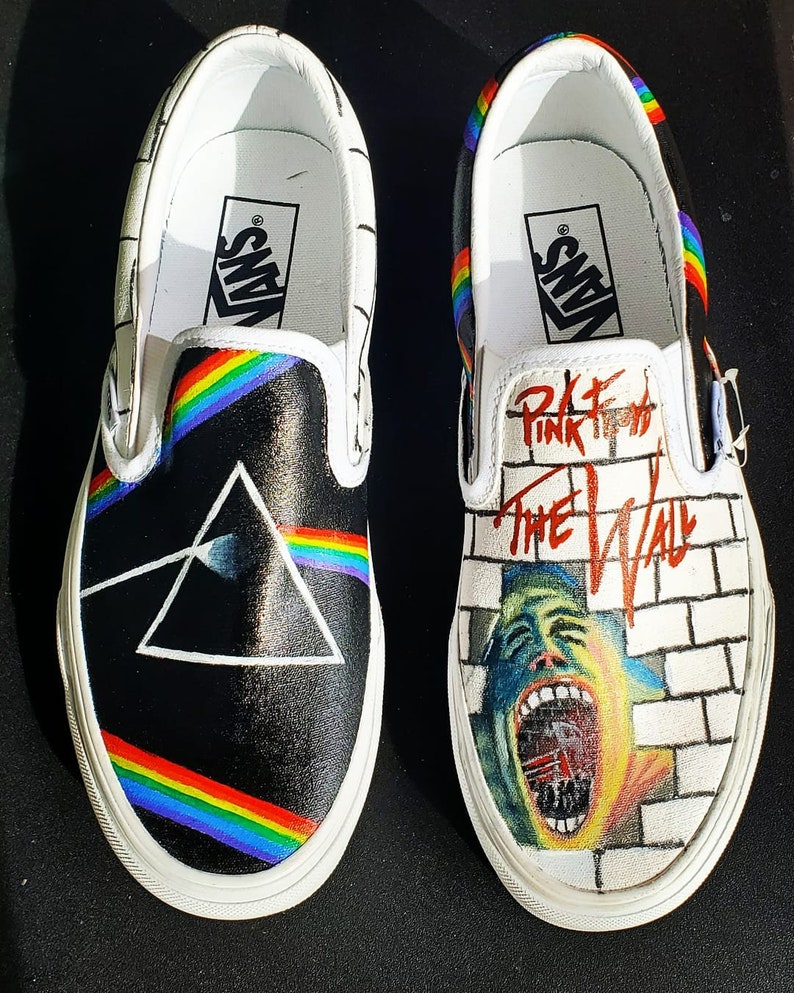 May include: A pair of white Vans slip-on shoes with custom artwork. One shoe features a black background with a rainbow and a prism, while the other shoe features a white brick wall with a red and black graphic of a man's face and the text "Pink Floyd The Wall".