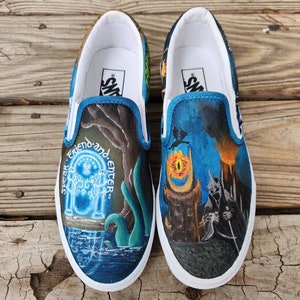 May include: A pair of blue and black Vans slip-on shoes with a hand-painted design featuring the Eye of Sauron and the inscription "Speak friend and enter."