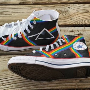 May include: A pair of black high-top Converse sneakers with a rainbow stripe design and a white triangle logo. The sneakers have white laces and a white rubber sole.
