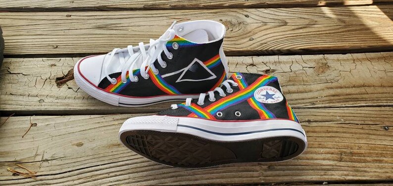 May include: A pair of black high-top Converse sneakers with a rainbow stripe design and a white triangle logo. The sneakers have white laces and a white rubber sole.