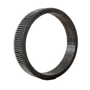 May include: A black circular gear with a toothed outer edge. The gear is made of a dark material and is set against a white background. The teeth are evenly spaced around the circumference, creating a textured appearance.