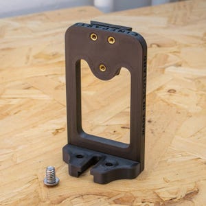 Może przedstawiać: Czarny plastikowy uchwyt na telefon z prostokątnym otworem i czterema złotymi śrubami. Uchwyt ma nadrukowany tekst "MADE IN SCOTLAND" na boku. Srebrna śruba leży na drewnianej powierzchni.