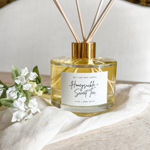 May include: A clear glass bottle with a gold lid and wooden sticks, filled with a light yellow liquid. The bottle has a white label with the text "SALT + LIGHT BATH COMPANY" and "Honeysuckle + Sweet Tea" in black script. The bottle is sitting on a white surface with a white flower.