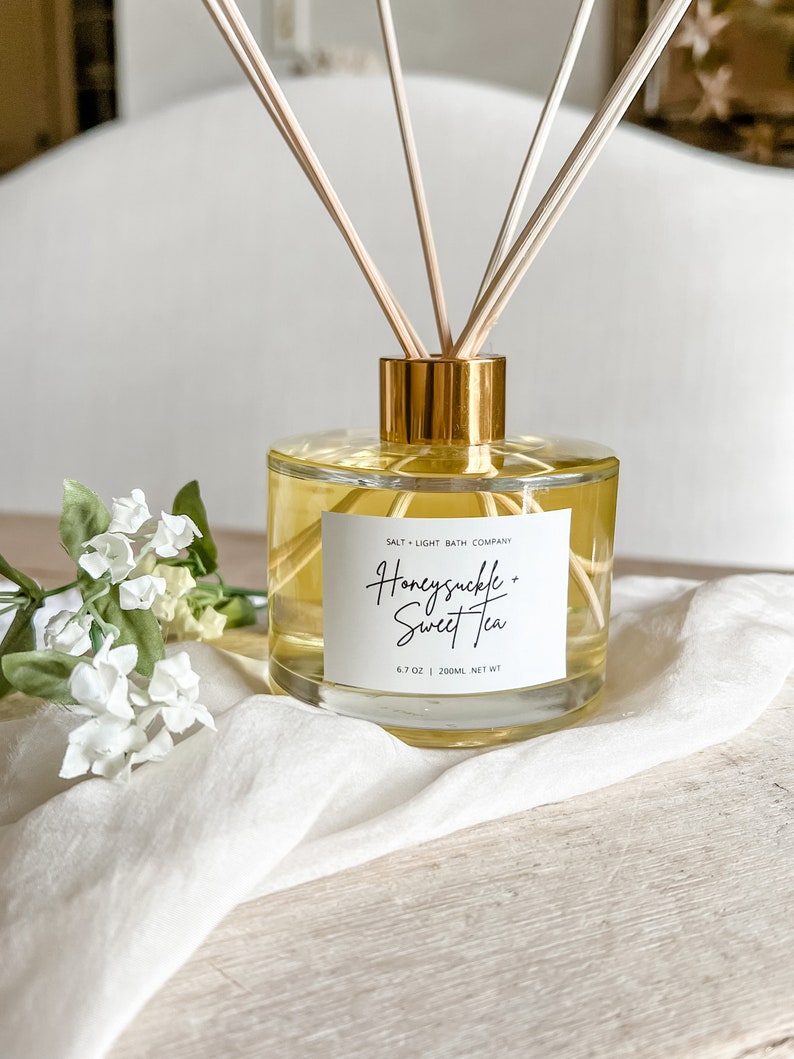 May include: A clear glass bottle with a gold lid and wooden sticks, filled with a light yellow liquid. The bottle has a white label with the text "SALT + LIGHT BATH COMPANY" and "Honeysuckle + Sweet Tea" in black script. The bottle is sitting on a white surface with a white flower.