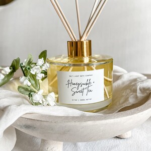 May include: A clear glass bottle with a gold lid and wooden sticks, filled with a yellow liquid. The bottle has a white label that reads "Salt + Light Bath Company" and "Honeysuckle + Sweet Tea" with a 6.7 oz | 200ml net wt label. The bottle is sitting on a white wooden tray with three legs.