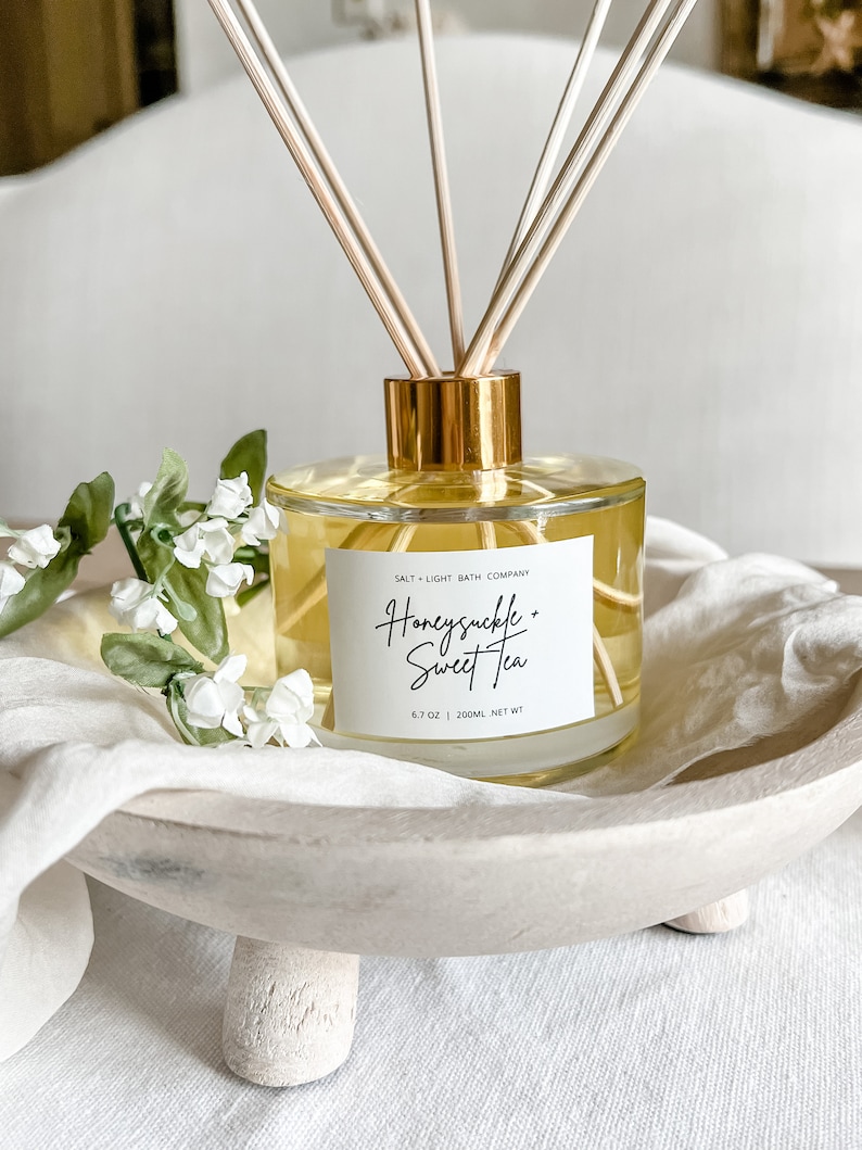 May include: A clear glass bottle with a gold lid and wooden sticks, filled with a yellow liquid. The bottle has a white label that reads "Salt + Light Bath Company" and "Honeysuckle + Sweet Tea" with a 6.7 oz | 200ml net wt label. The bottle is sitting on a white wooden tray with three legs.
