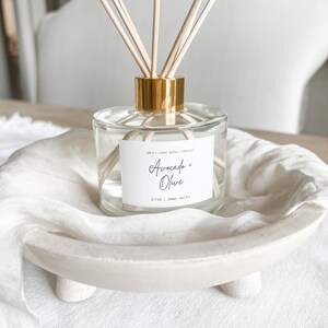 K&ouml;nnte beinhalten: Eine transparente Glasdiffusorflasche mit goldenem Verschluss und Holzst&auml;bchen. Das Etikett tr&auml;gt die Aufschrift "Avocado + Olive" von Salt + Light Bath Company. Die Flasche steht in einer wei&szlig;en, flachen Holzschale auf einer wei&szlig;en Leinenoberfl&auml;che.