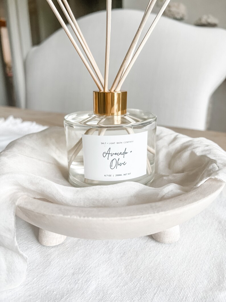 K&ouml;nnte beinhalten: Eine transparente Glasdiffusorflasche mit goldenem Verschluss und Holzst&auml;bchen. Das Etikett tr&auml;gt die Aufschrift "Avocado + Olive" von Salt + Light Bath Company. Die Flasche steht in einer wei&szlig;en, flachen Holzschale auf einer wei&szlig;en Leinenoberfl&auml;che.